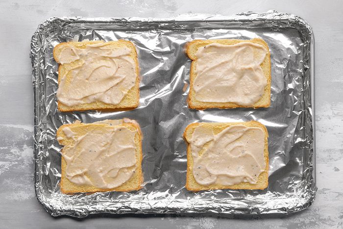 Four slices of bread topped with a creamy spread are arranged on a foil-lined baking sheet, ready to be baked.