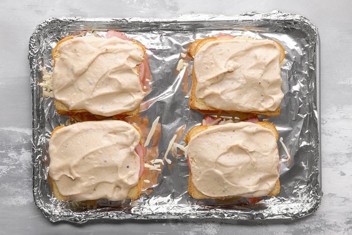 Four open-faced sandwiches topped with a creamy sauce are arranged on a foil-lined baking sheet, ready to be baked. The bread, filling, and sauce are visible, and the surface beneath the tray is light gray.