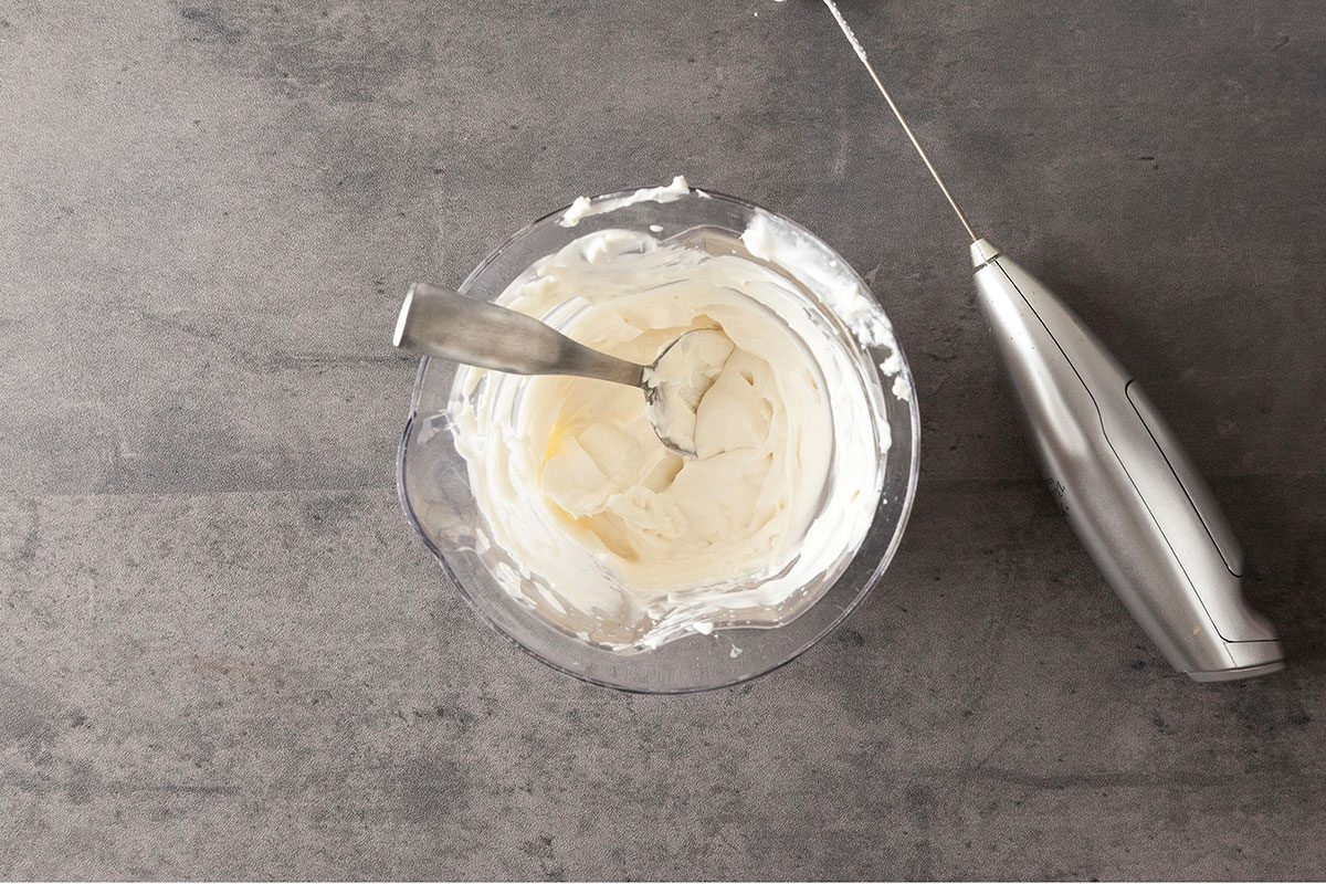 A bowl of whipped cream with a spoon inside sits on a gray surface next to a silver handheld electric mixer.