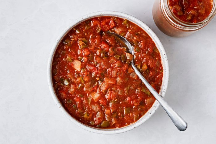 A bowl of chunky tomato salsa with diced vegetables and a spoon, next to a jar filled with the same salsa, on a light-colored surface.