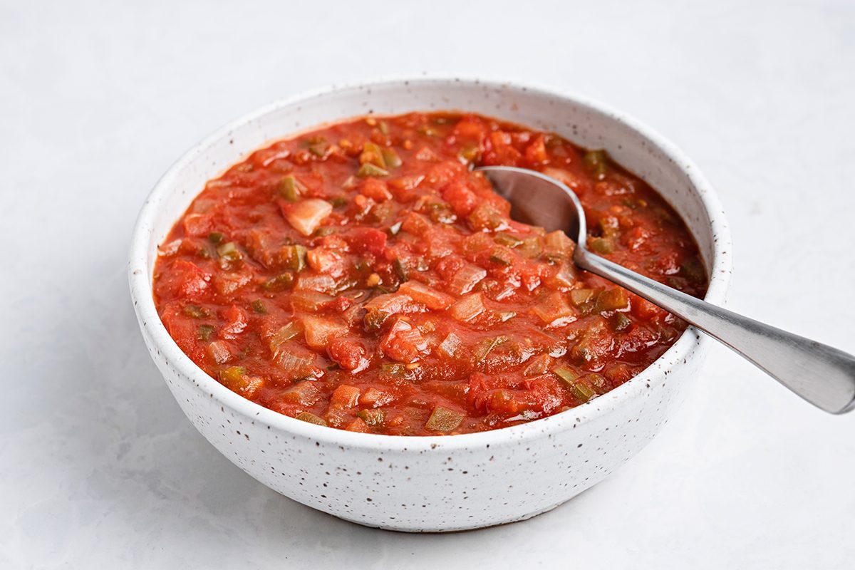 A white speckled bowl filled with chunky red salsa, containing visible pieces of tomato, onion, and peppers, with a metal spoon resting inside the bowl on a light surface.