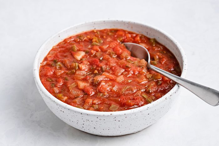 A white speckled bowl filled with chunky red salsa, containing visible pieces of tomato, onion, and peppers, with a metal spoon resting inside the bowl on a light surface.
