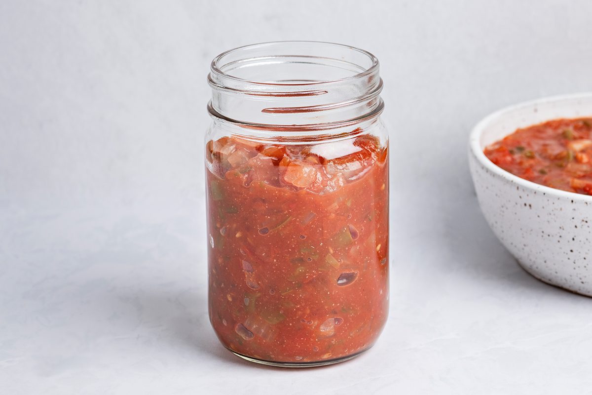 A glass mason jar filled with chunky red salsa sits on a light surface. In the background, there is a white bowl containing more salsa.