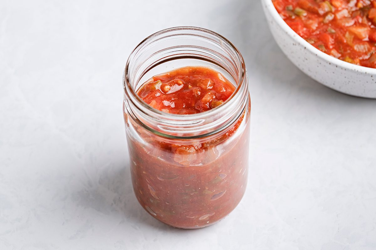 A glass jar filled with chunky red salsa sits on a light gray surface. Part of a white bowl containing more salsa is visible in the background.
