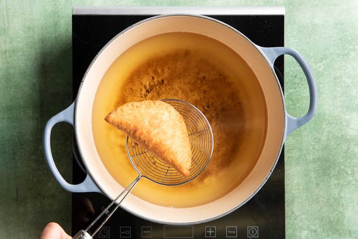 Taking out a fried apple pie from a frying pan kept on top of a black induction cooktop on a green marble countertop.
