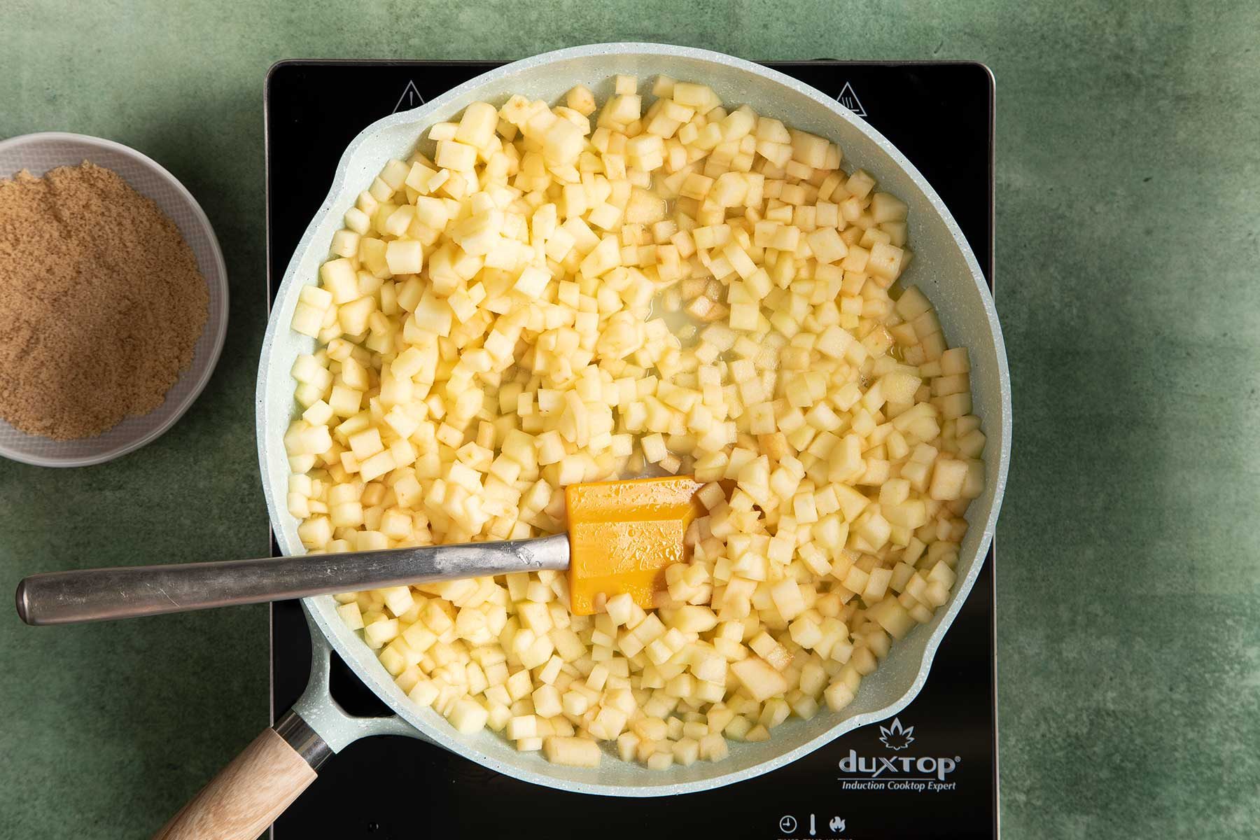 Overhead shot of cooking the apple filling in ceramic cookware on a black induction top kept on top of a green marble countertop, with brown sugar kept beside it.