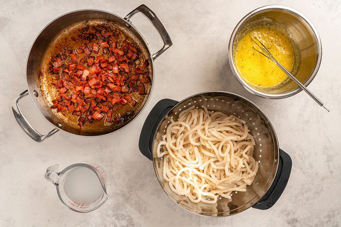 Crisp bacon in a saucepan, boiled udon noodles in pan and beated eggs in small pan placed on table