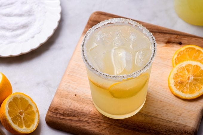 High angle shot of a glass of light yellow iced drink with lemon wedges on top of a cutting board, next to a plate of salt