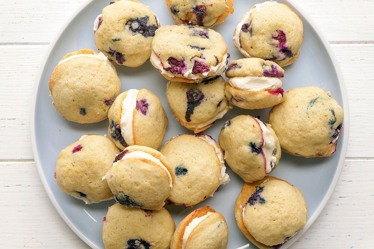 overhead shot of Several lemon blueberry whoopie pies are arranged on a light blue plate, the plate rests on a white wooden surface