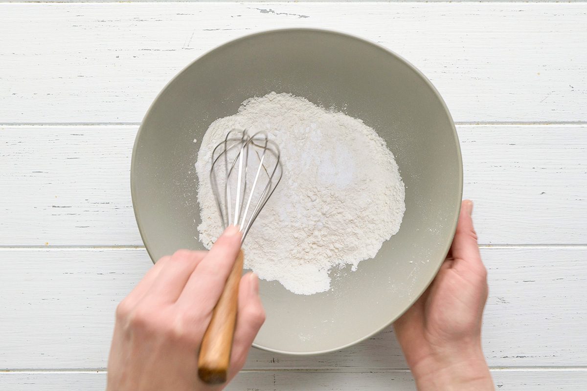 overhead shot of Two hands are holding a gray bowl containing a mixture of flour and baking powder or salt; a whisk with a wooden handle is partially submerged in the mixture, the bowl is positioned over a white wooden surface