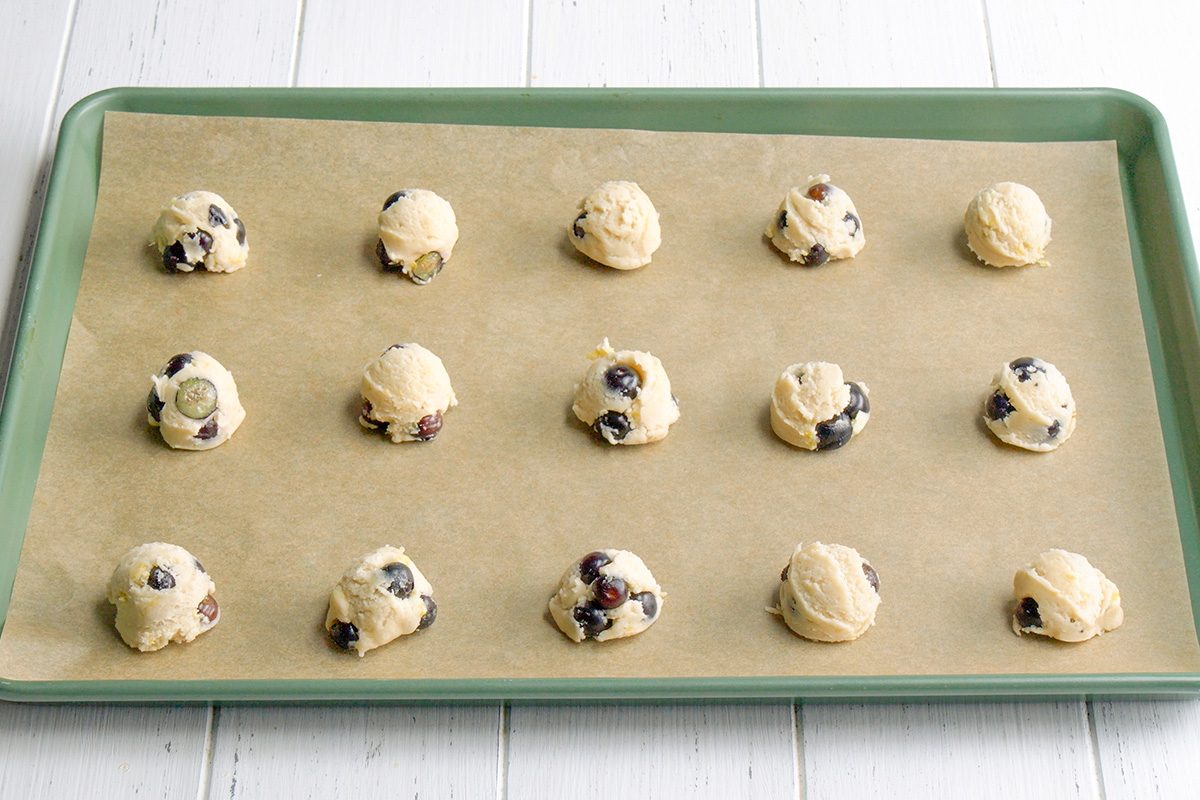 3/4th shot of Fifteen pale, round balls of cookie dough, dotted with blueberries, are arranged on a rectangular baking sheet lined with parchment paper; the baking sheet has a dark green rim and rests on a white wooden surface