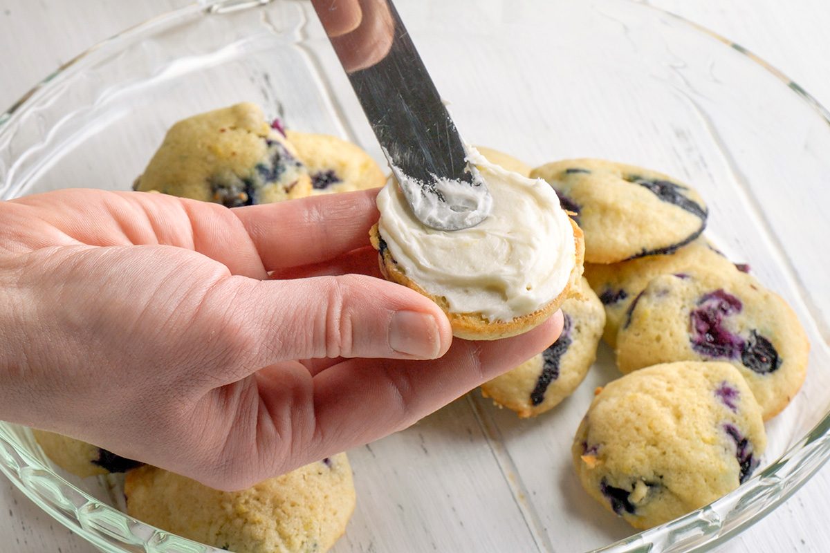 3/4th shot of A hand is holding a small, round, pale yellow cookie dotted with blueberries, and a metal spatula is spreading a thick layer of creamy white frosting on its flat surface; several other similar cookies are visible in a clear glass dish below, the dish rests on a white wooden surface