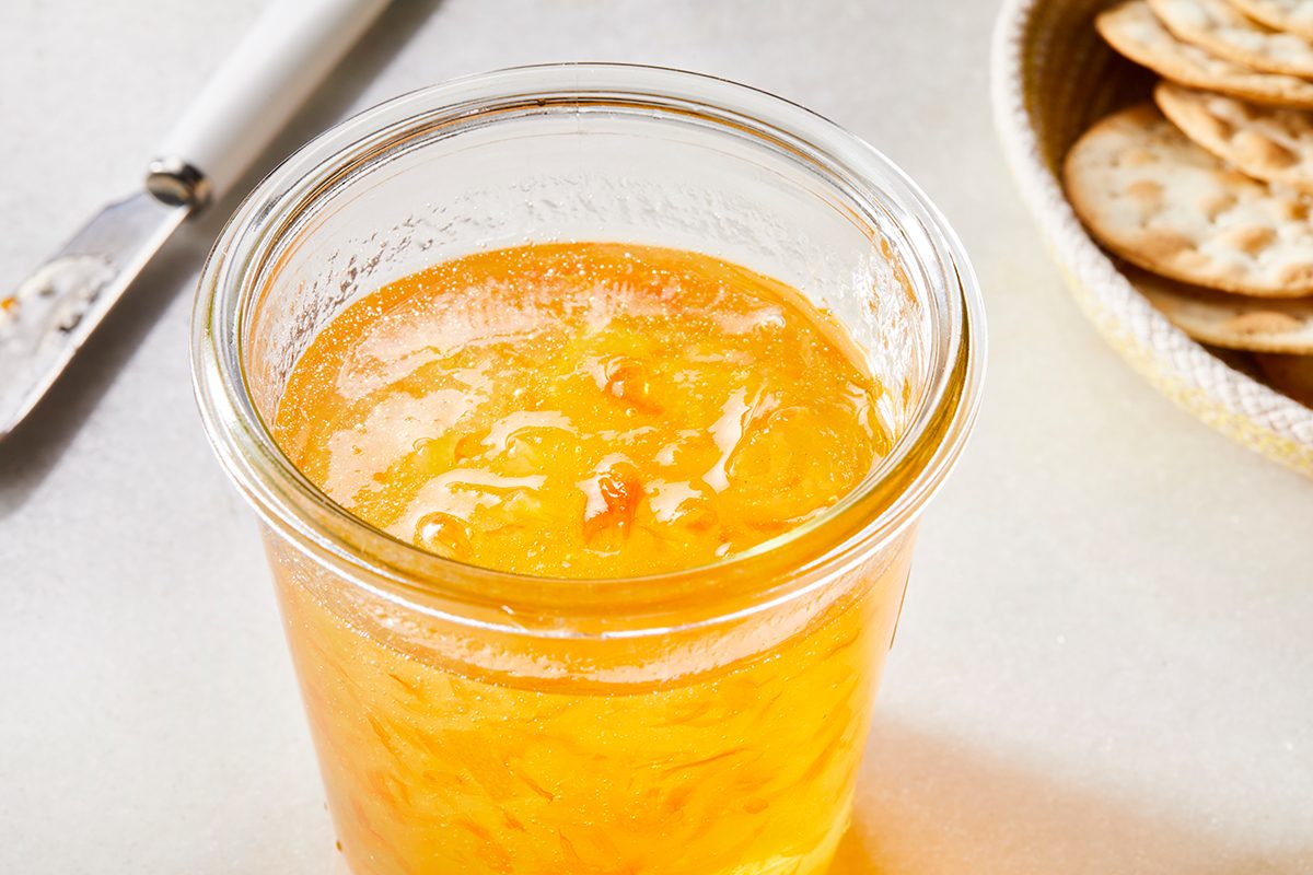 A glass jar filled with orange marmalade sits on a light surface, with a butter knife and a plate of round crackers in the background.