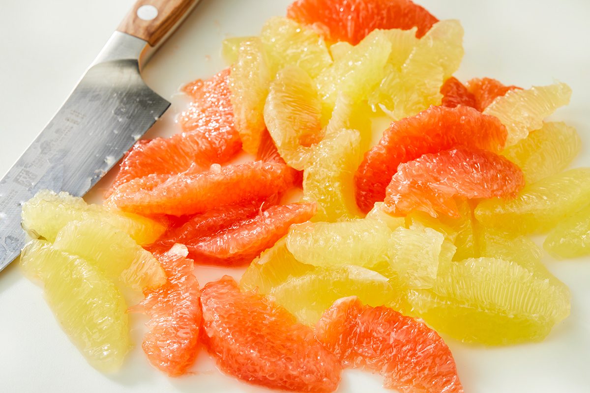 Segments of pink and yellow citrus fruit are arranged on a white surface next to a knife with a wooden handle.