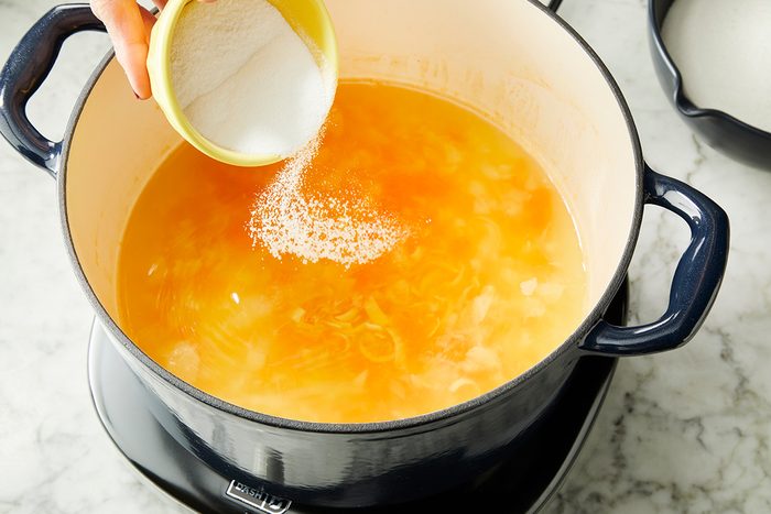 A hand pours salt from a small bowl into a large pot of yellow-orange liquid, possibly soup or broth, on a stovetop.