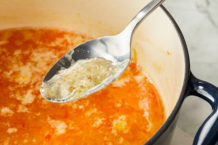 A metal spoon skims off foamy scum from the surface of an orange-colored soup or broth cooking in a large pot on a stovetop.