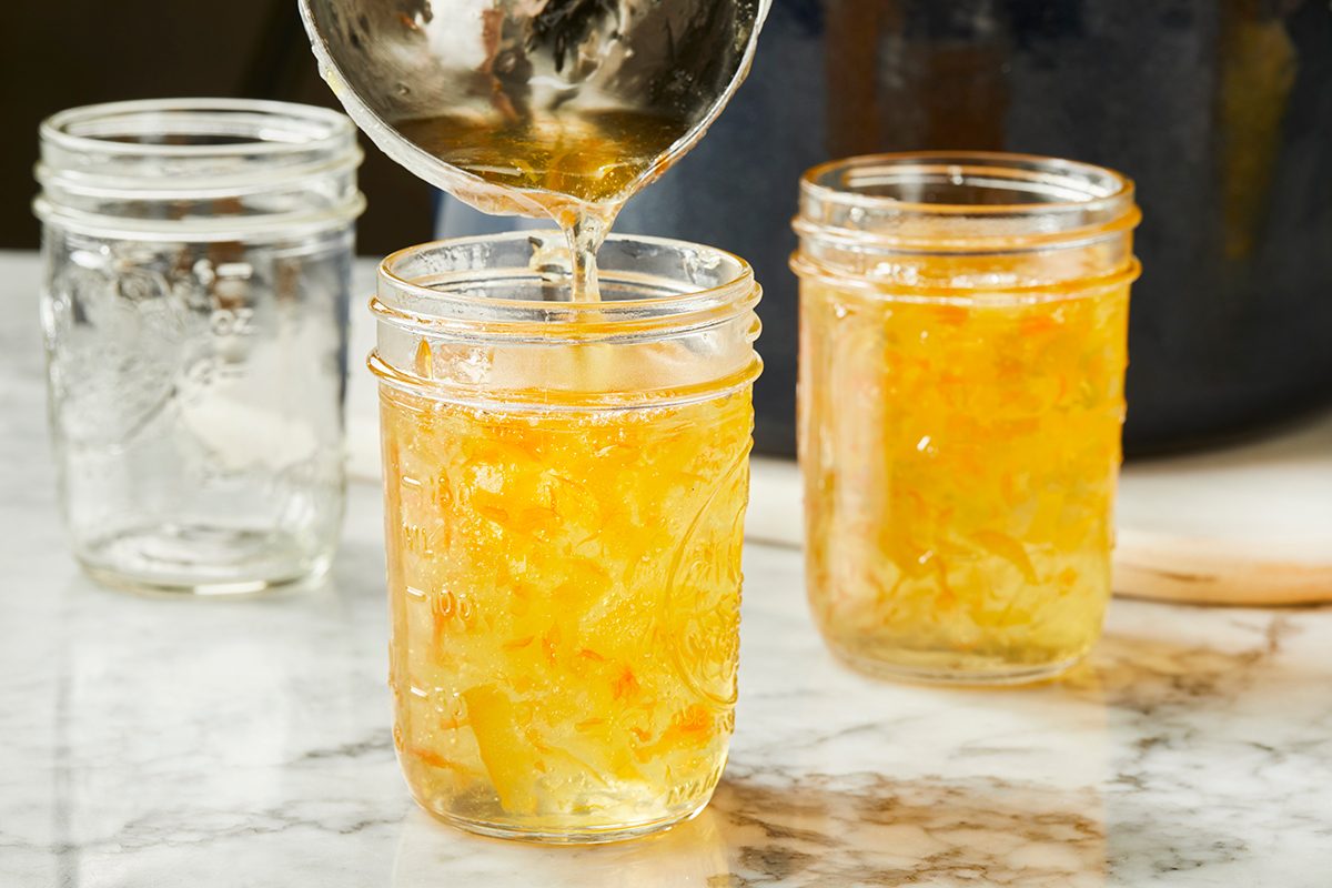 A close-up of marmalade being poured from a ladle into a glass jar on a marble countertop, with two jars—one filled and one empty—visible in the background.