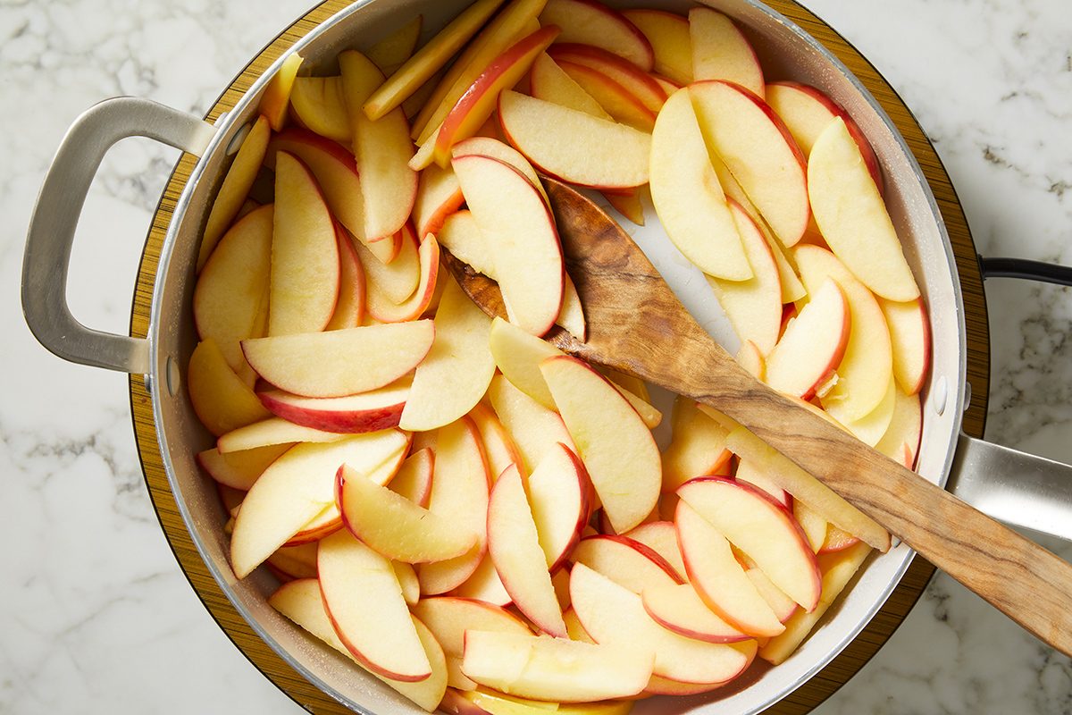 Sliced red and yellow apples in a pot, being stirred with a wooden spoon, on a marble countertop.