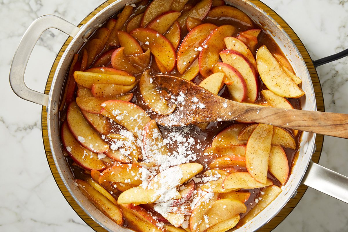 Sliced apples being stirred with a wooden spoon in a pot, mixed with brown sugar, spices, and a sprinkle of cornstarch, on a white marble surface.