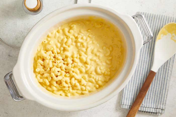 overhead shot of a dish of Potluck Macaroni and Cheese presented in a glossy white serving dish; beside the dish, there is a spoon with a wooden handle resting on a light blue and white striped napkin, the background is a light, neutral surface