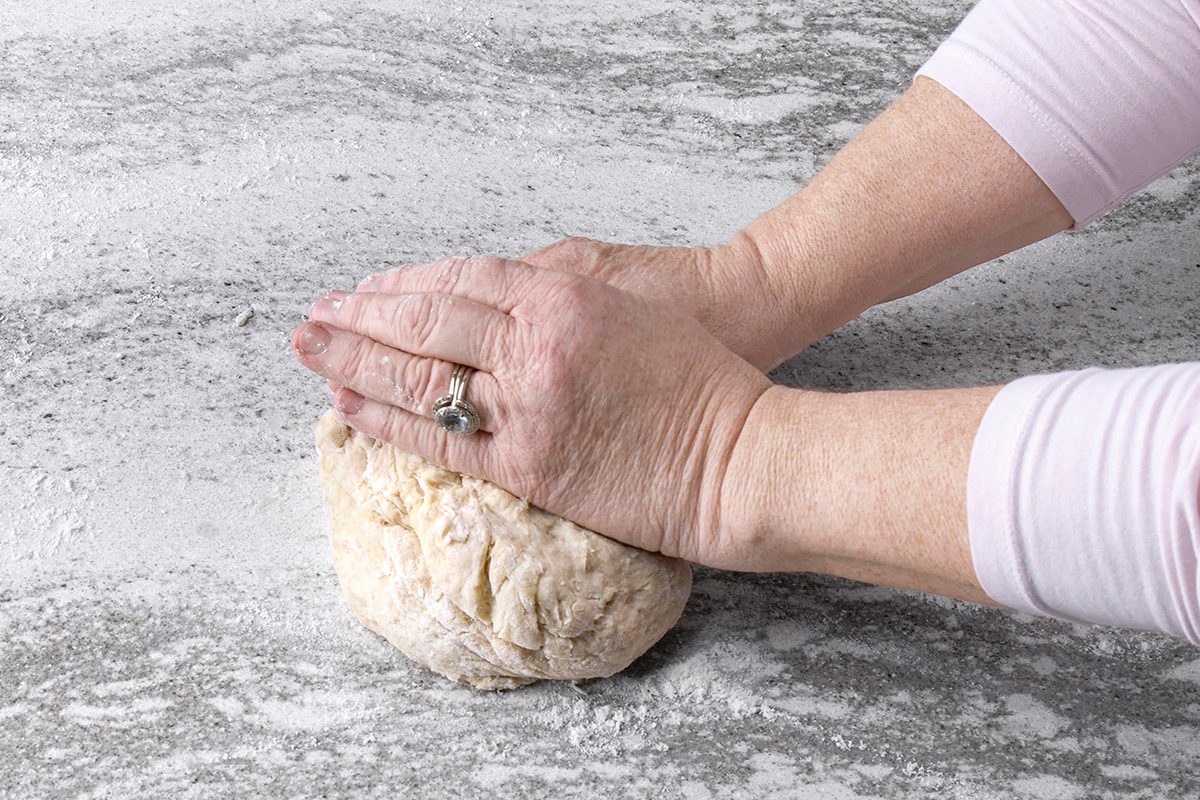 Hands kneading a ball of dough on a floured, gray countertop.