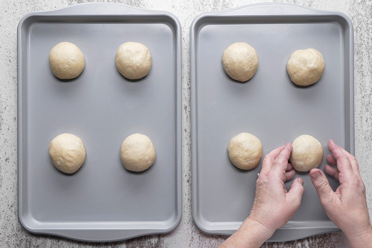 Two baking sheets with three evenly spaced dough balls on each. A pair of hands is shaping one dough ball on the right tray, preparing the dough on a light-colored surface.