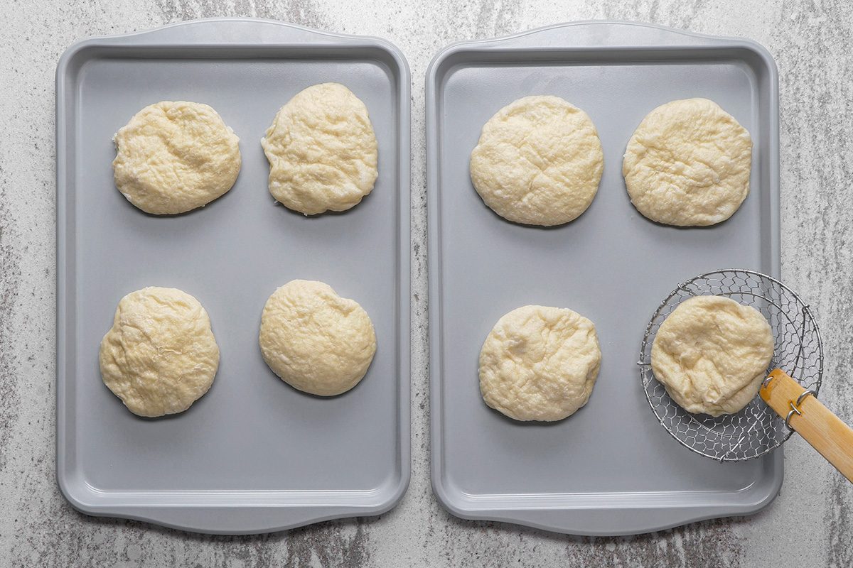 Two baking sheets each hold three round, uncooked dough balls. A slotted spoon holds one dough ball above the right tray, on a gray countertop.