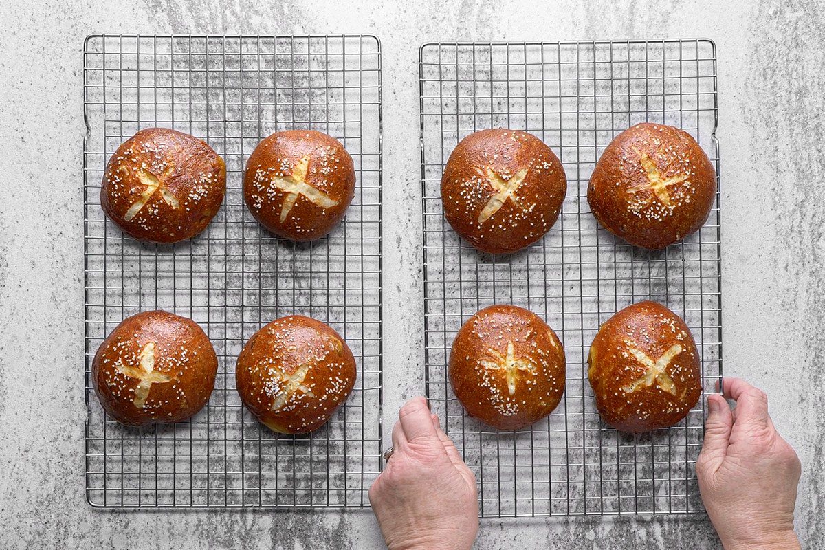 Two cooling racks, each holding four round pretzel buns topped with salt and scored with an X, are placed on a gray countertop. Hands are holding the rack on the right.