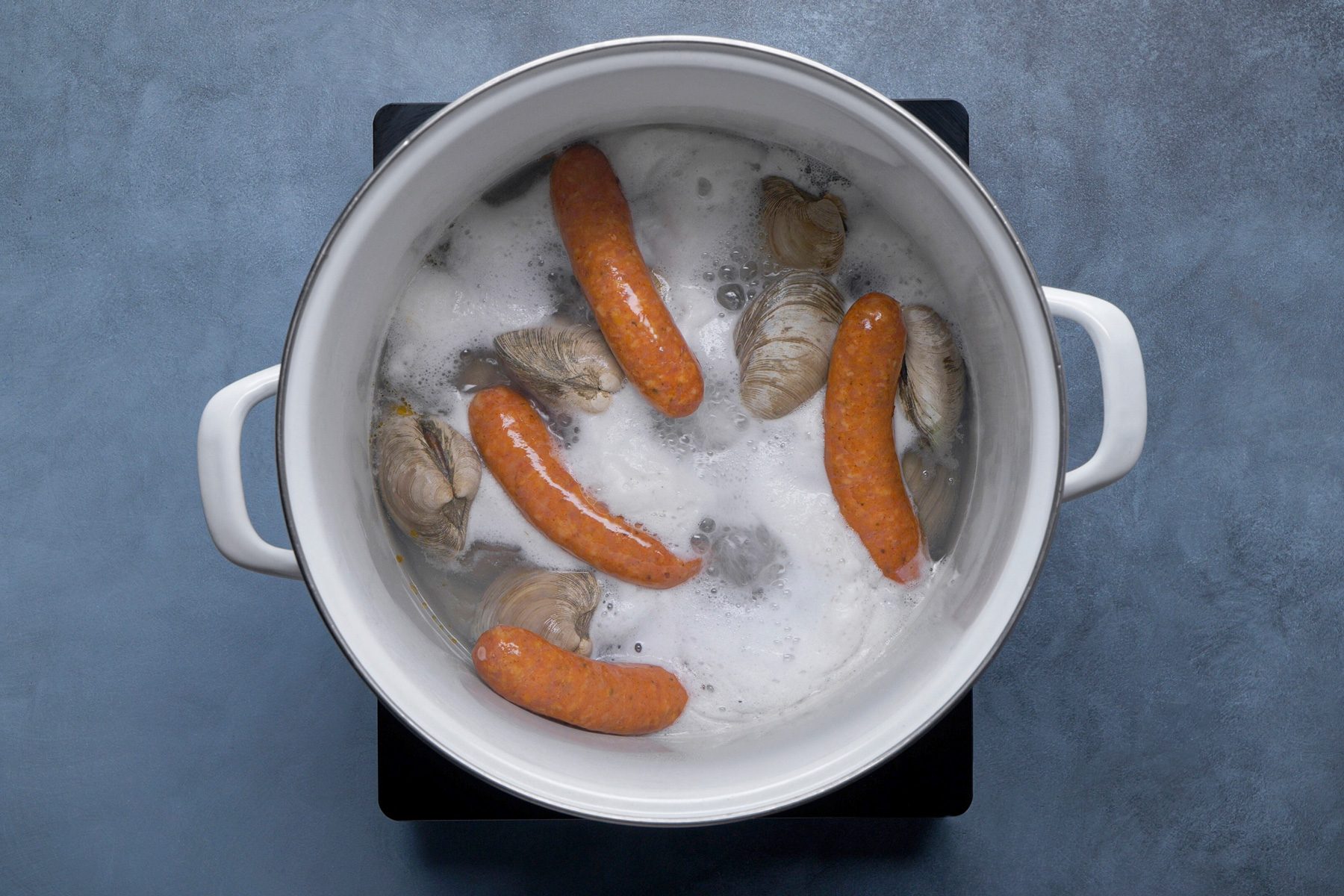 overhead shot of a white pot filled with boiling water, sausages and several open clam shells; the sausages are submerged in the bubbling water, and the clam shells are scattered around them; the pot has white handles and is sitting on a black electric stovetop; the background is a blue gray surface