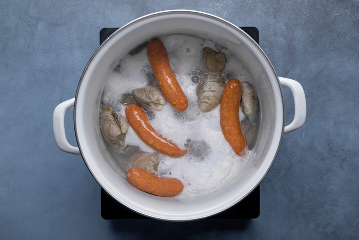 overhead shot of a white pot filled with boiling water, sausages and several open clam shells; the sausages are submerged in the bubbling water, and the clam shells are scattered around them; the pot has white handles and is sitting on a black electric stovetop; the background is a blue gray surface