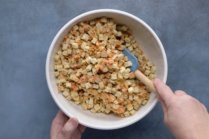 overhead shot of Two hands are holding a white bowl filled with a mixture of small, light brown bread cubes, possibly unseasoned stuffing cubes, and some darker brown flecks, a blue silicone spatula with a wooden handle is partially submerged in the mixture, the bowl is positioned over a solid, muted blue gray surface