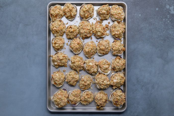overhead shot of a silver baking sheet filled with numerous stuffed clam shells, arranged in neat rows; each clam shell is filled with a light brown, breadcrumb based stuffing, likely containing chopped seafood and vegetables; the baking sheet has a slightly raised rim and rests on a solid, muted blue gray surface