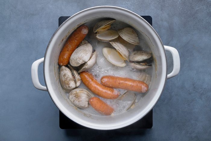 overhead shot of a white pot filled with boiling water, sausages and several open clam shells; the sausages are submerged in the bubbling water, and the clam shells are scattered around them; the pot has white handles and is sitting on a black electric stovetop; the background is a blue gray surface