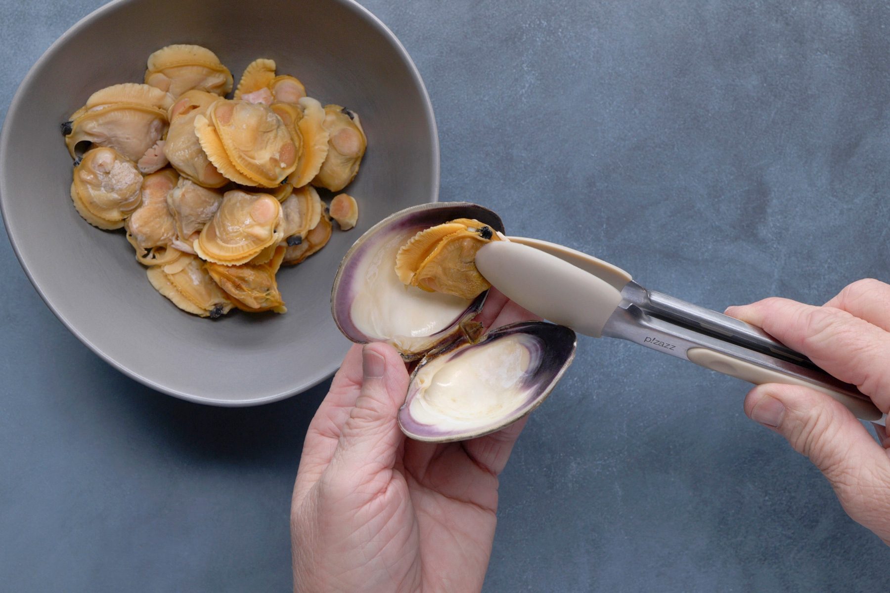 Two hands are shown, one holding an open clam shell and the other using tongs to remove the clam meat; a bowl filled with cooked clams is visible in the background, positioned to the left of the open clam; the background is a solid gray surface