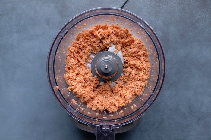overhead shot of the inside of a clear plastic food processor bowl containing a chunky, light brown mixture, possibly homemade peanut butter or a similar nut butter; the bowl is sitting on the base of the food processor, which is partially visible at the bottom of the frame, and the background is a solid, muted blue gray surface