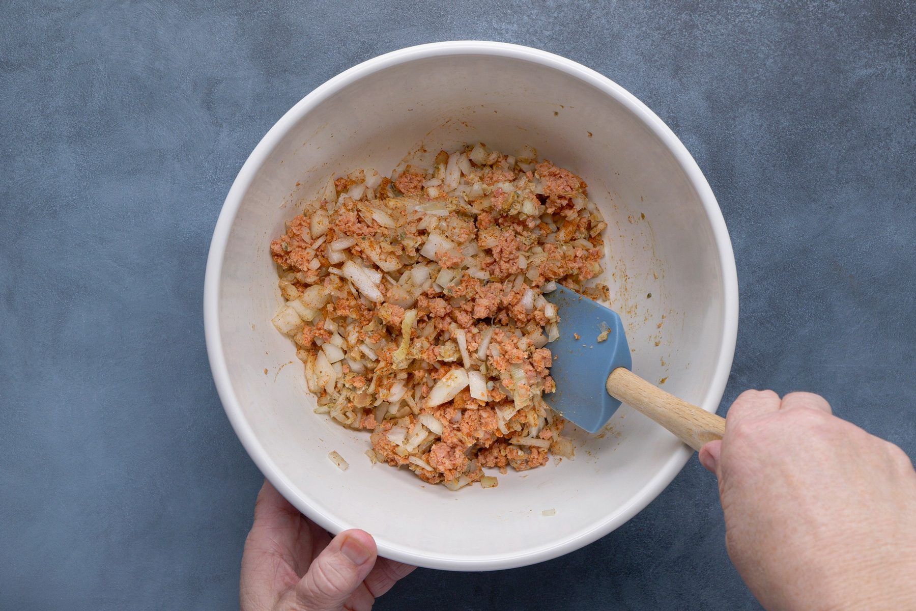 overhead shot of Two hands are holding a white bowl containing a mixture of finely chopped light pink meat, possibly canned tuna or shredded chicken, and translucent white pieces of onion or celery, all coated in a light brown seasoning or sauce; a blue silicone spatula with a wooden handle is partially submerged in the mixture, the bowl is positioned over a solid, muted blue gray surface
