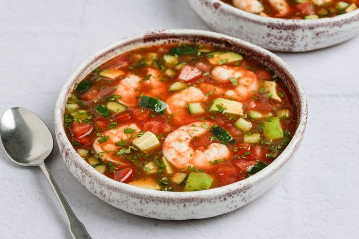 overhead shot of a dish called Shrimp Gazpacho; in two shallow bowls, next to the bowls are two shiny silver spoon, the background is a soft, light gray