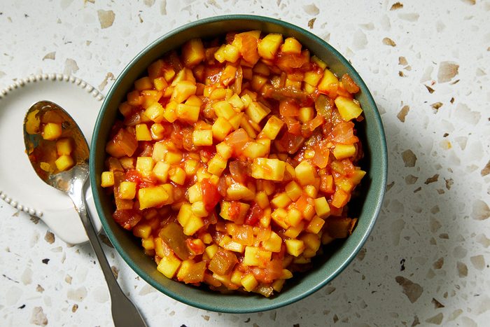 A bowl filled with a chunky fruit salsa made of diced mango, pineapple, and other colorful fruits sits on a speckled countertop. A spoon with some of the salsa rests beside the bowl.