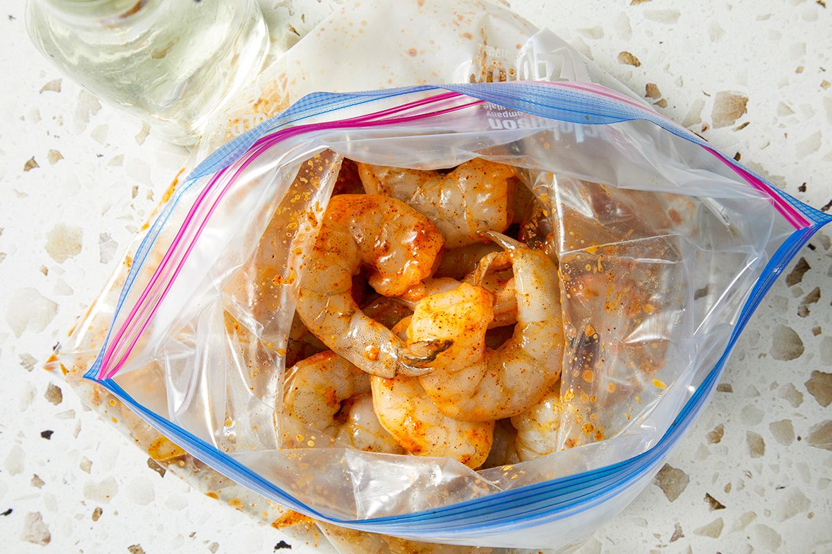 Raw shrimp marinated with spices inside a partially opened resealable plastic bag, placed on a white terrazzo countertop.