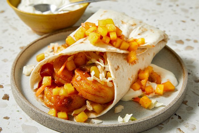 A tortilla wrap filled with cooked shrimp, shredded cabbage, and topped with diced mango salsa, served on a round plate with a bowl of sauce in the background.