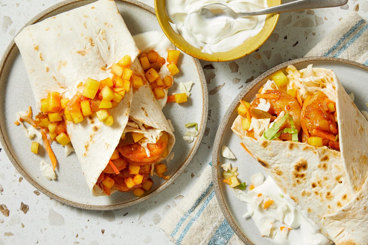 Two flour tortillas filled with shrimp, diced mango salsa, and shredded lettuce on plates. A small bowl of sour cream with a spoon is nearby, and some filling has spilled onto the plates.