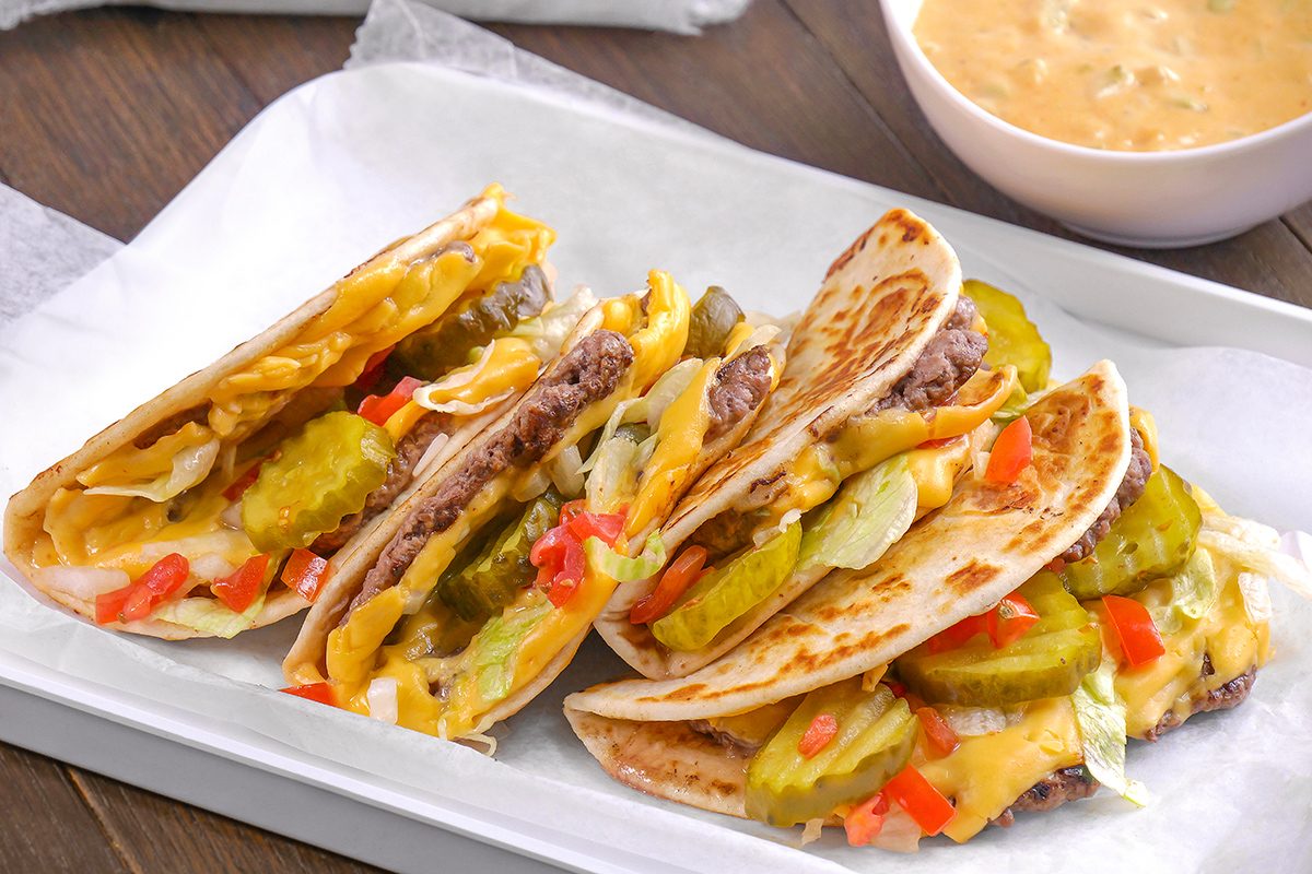 Three folded flour tortillas filled with ground beef, cheddar cheese, pickles, lettuce, and diced tomatoes are arranged on a white tray, with a bowl of dipping sauce in the background.