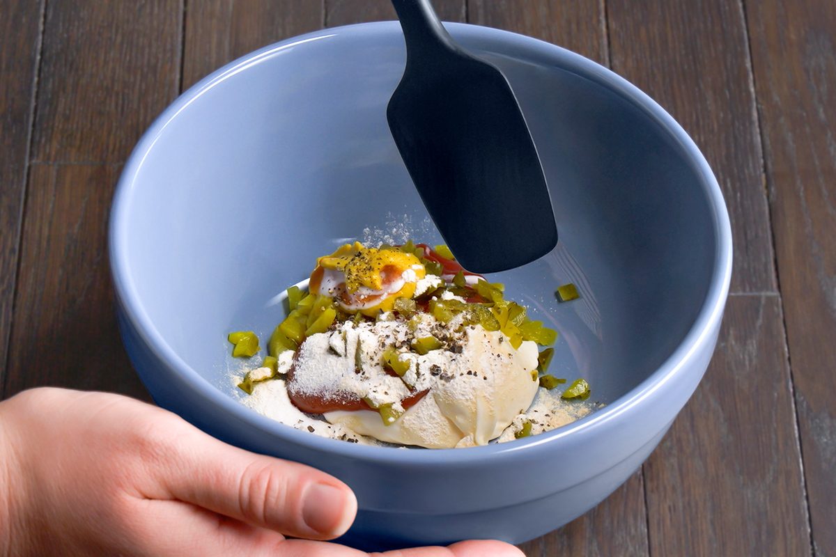 A hand holds a blue mixing bowl filled with various ingredients, including mayonnaise, mustard, and chopped pickles, while a spatula hovers above, ready to mix. The bowl is on a dark wooden surface.