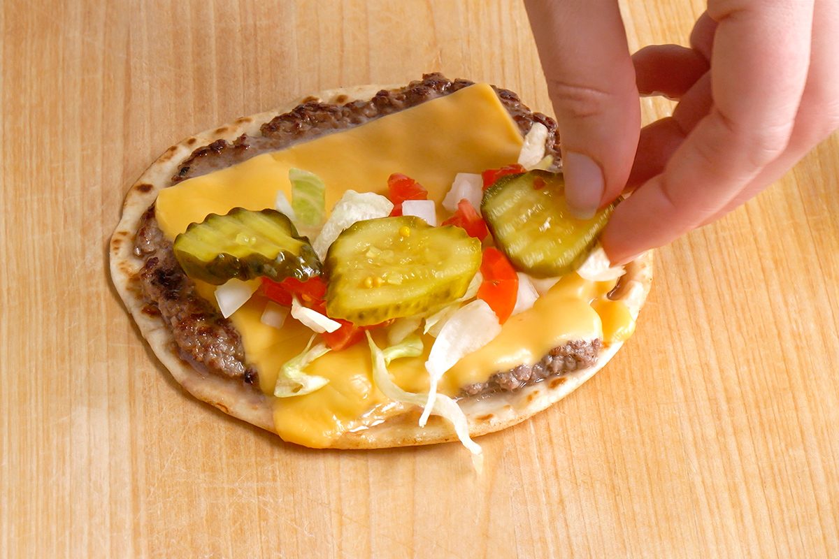A hand is placing pickle slices on a flatbread topped with ground beef, melted cheese, shredded lettuce, chopped onions, and diced tomatoes on a wooden surface.