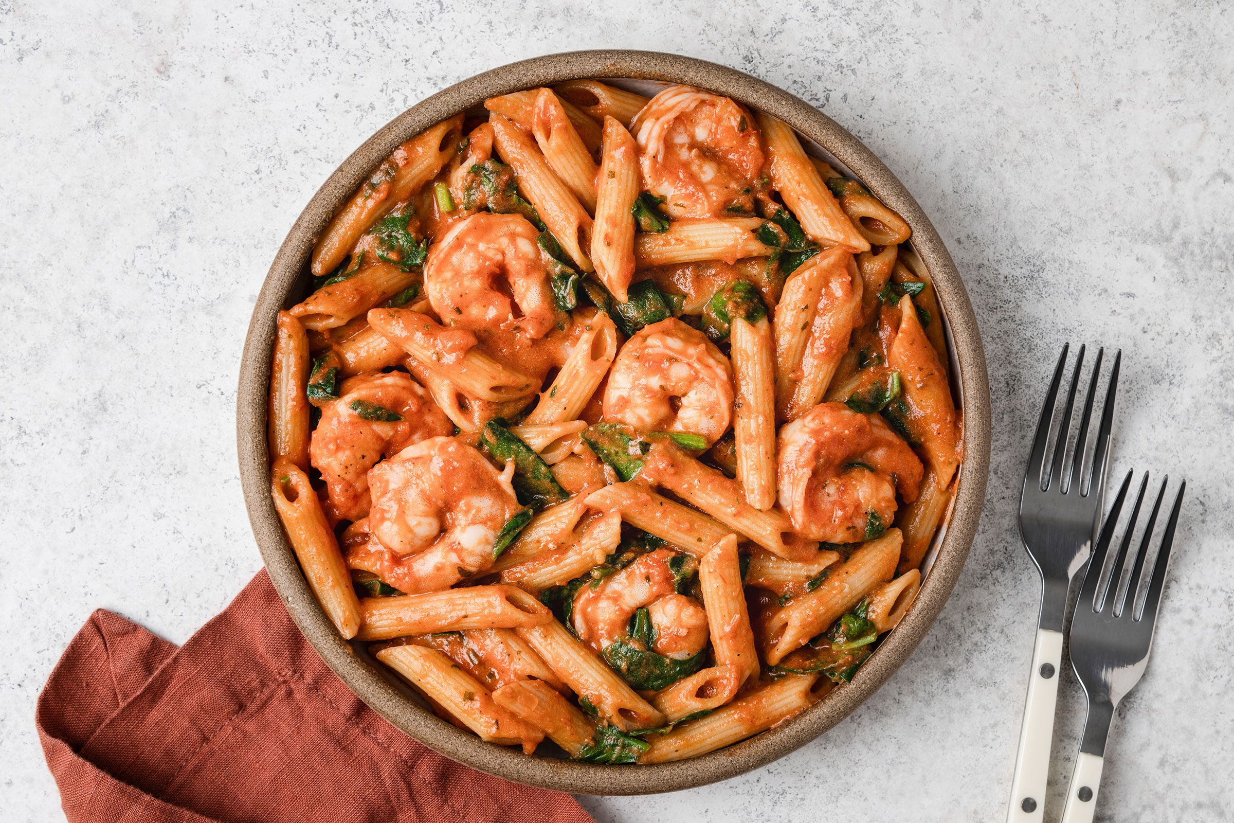 Spicy Shrimp And Penne Pasta with spinach in a bowl and some cutlery placed on side.