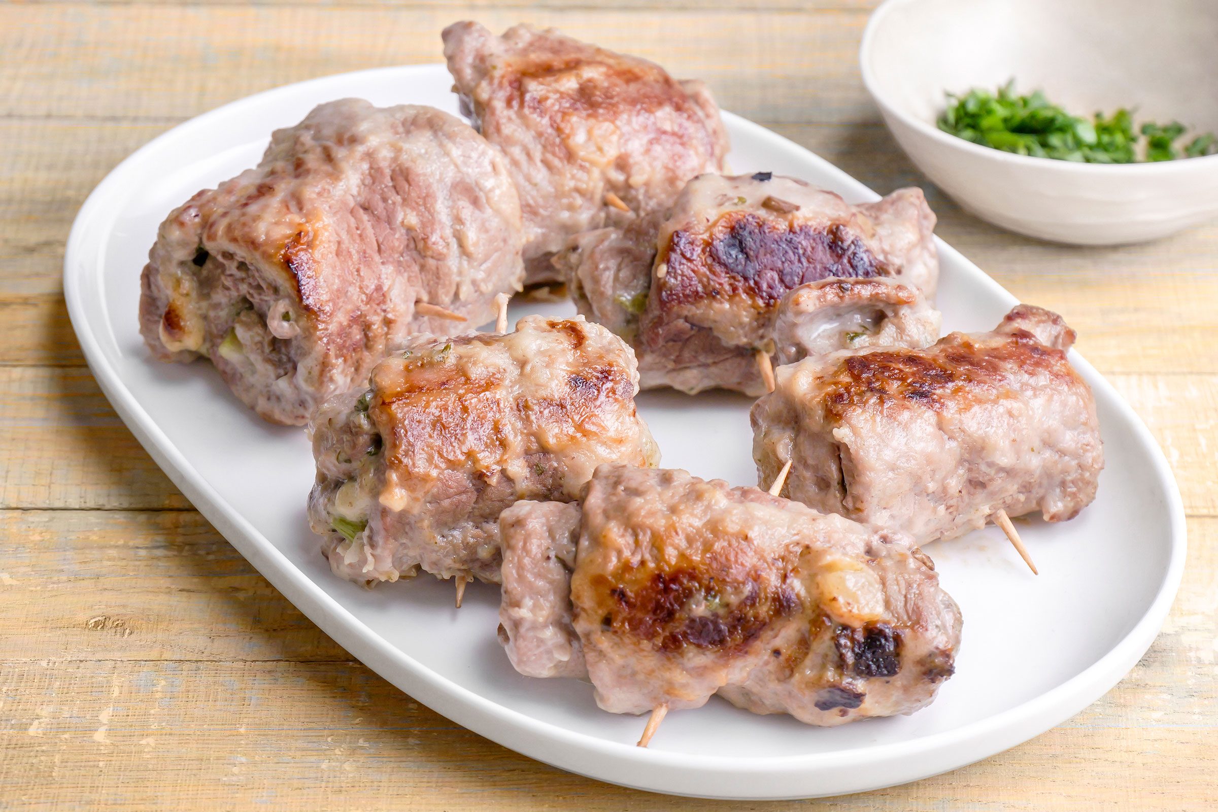 Steak Roll Ups served on a white serving tray with some green garnishing in a small bowl on side.