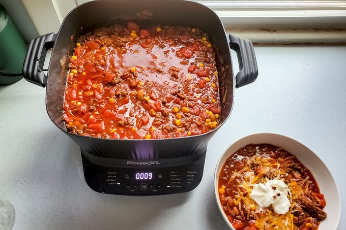 A pot of chili with ground meat, corn, beans, and tomatoes sits on an electric cooker next to a bowl of chili topped with shredded cheese and a dollop of sour cream.