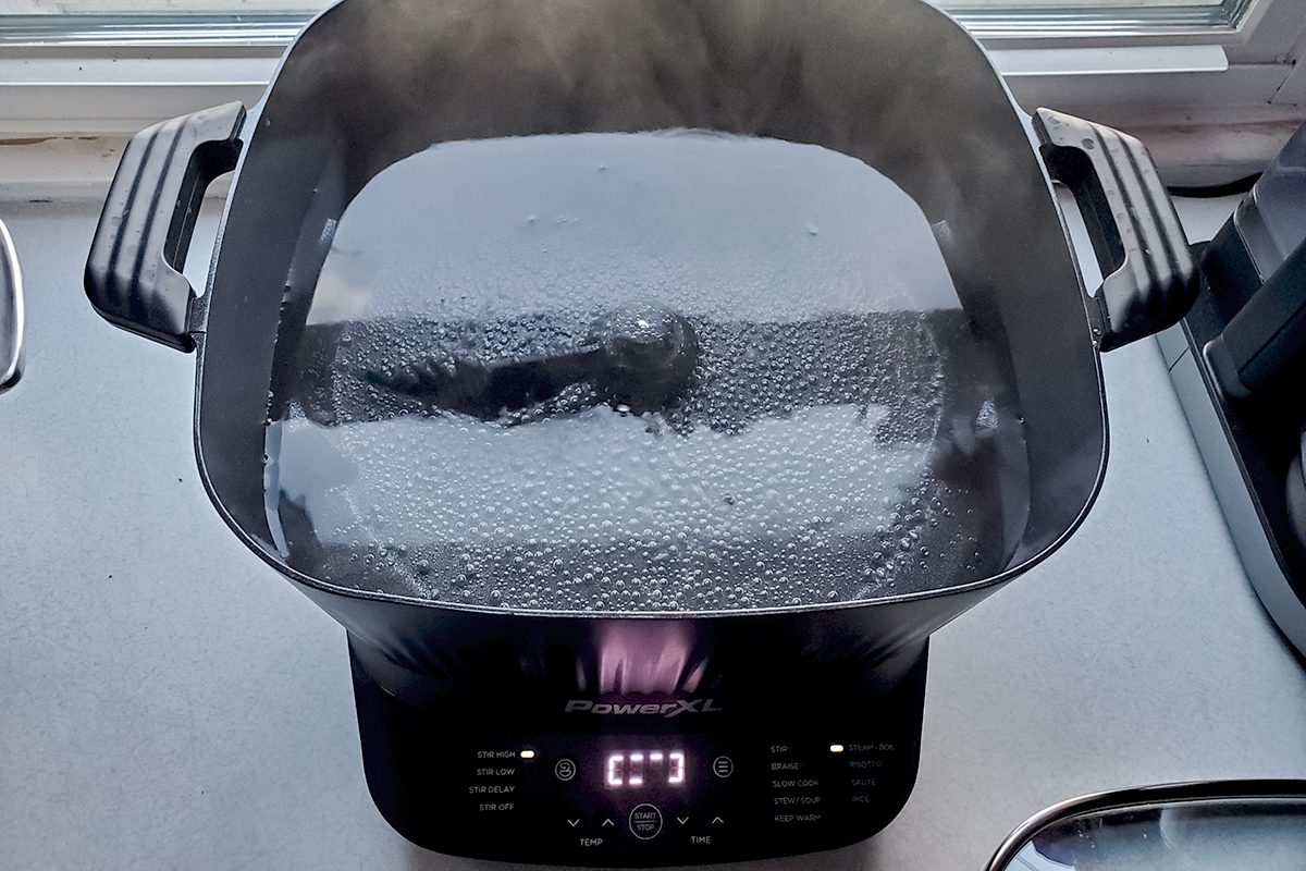A black electric cooker with a glass lid is steaming, and the digital display reads "0:17." The cooker is on a kitchen counter near a window.