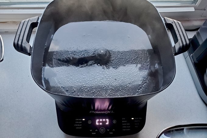 A black electric cooker with a glass lid is steaming, and the digital display reads "0:17." The cooker is on a kitchen counter near a window.