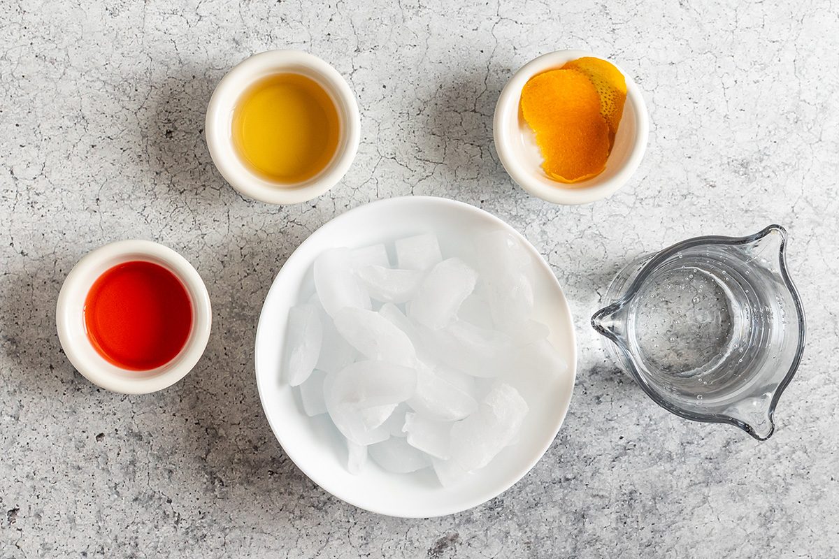A white bowl of ice cubes is surrounded by a small glass pitcher of water and three small bowls containing red, yellow, and orange liquids, all arranged on a light gray surface.